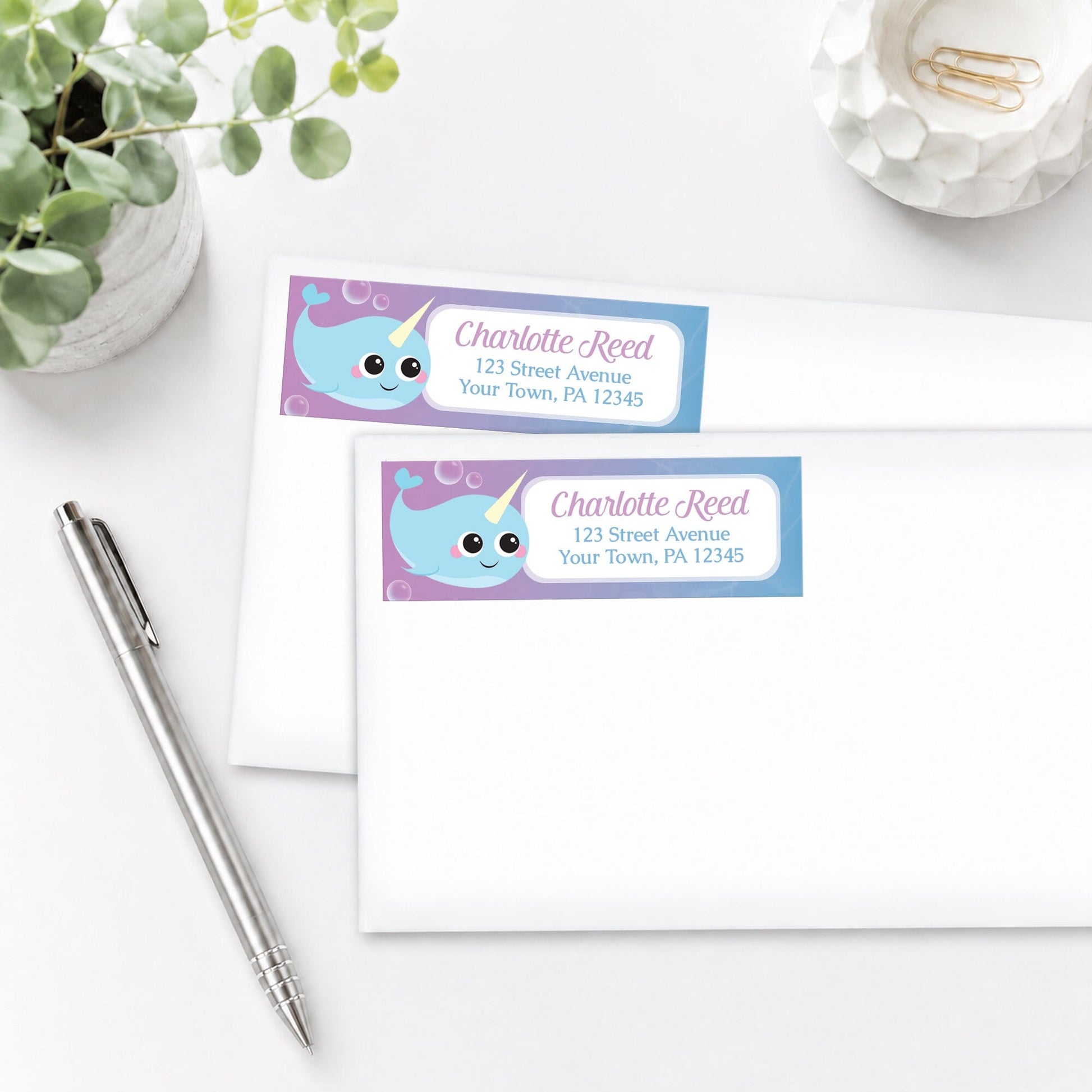 Cute Happy Narwhal Address Labels on white envelopes shown on a white surface with a plant and a pen.