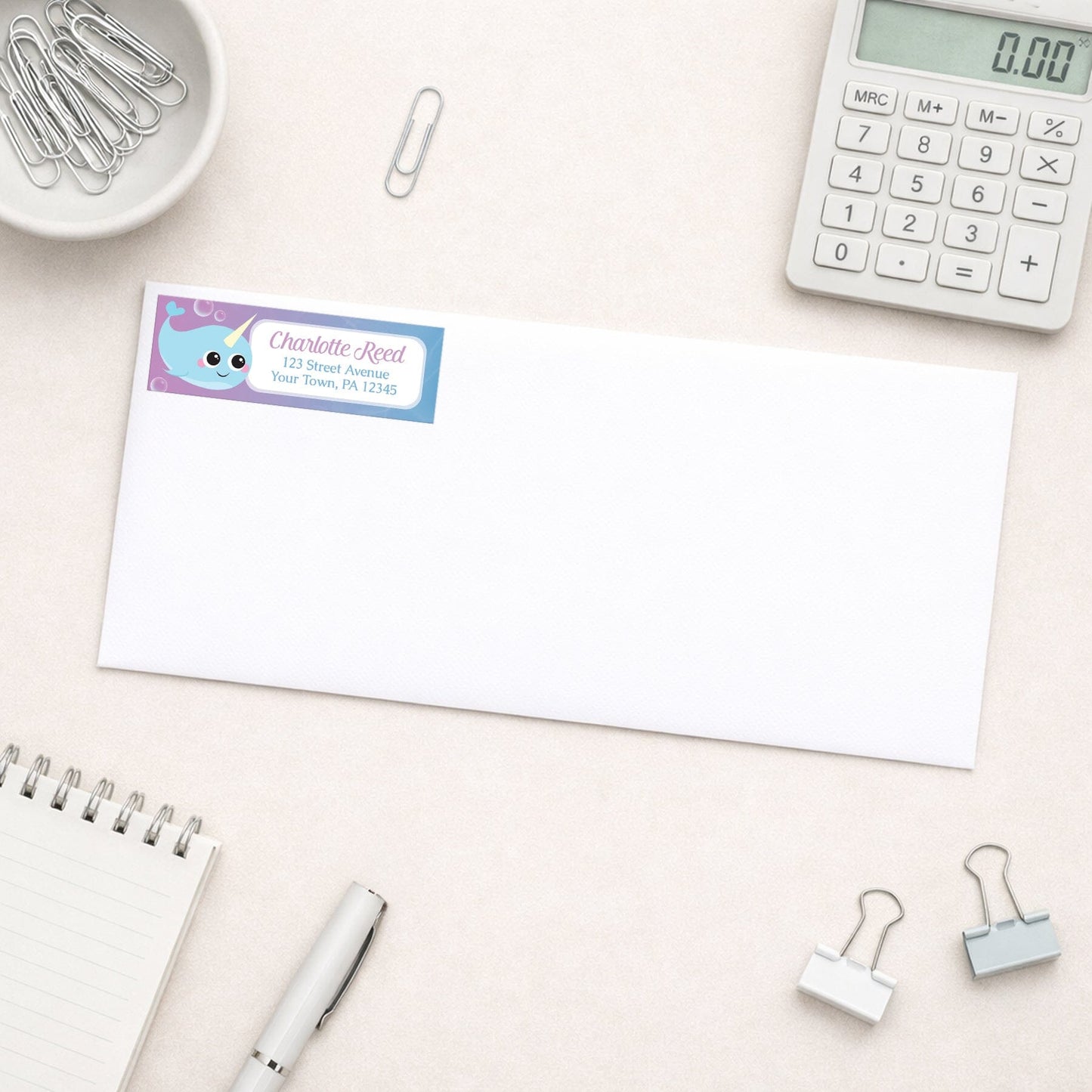 Cute Happy Narwhal Return Address Label shown on a white envelope with everyday office supplies