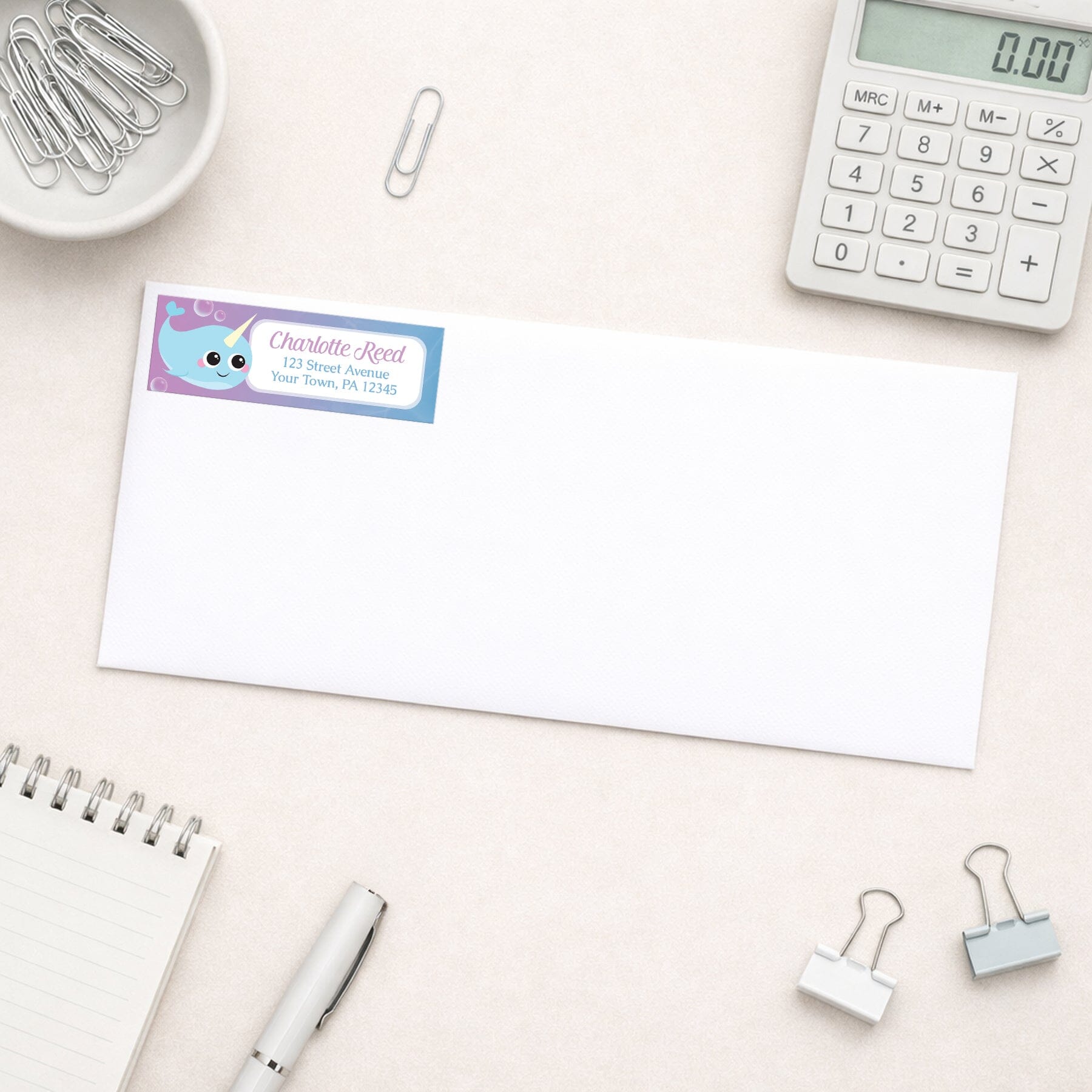 Cute Happy Narwhal Return Address Label shown on a white envelope with everyday office supplies