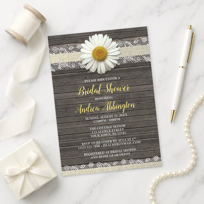 Bridal shower invitation with daisy and lace design on a wooden background, shown on a marble surface with decorative items.