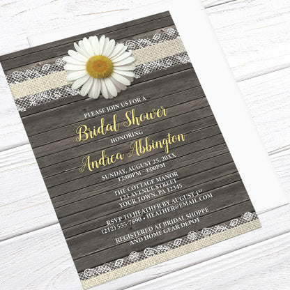 Floral bridal shower invitations with an illustrated design featuring a white dairy, burlap and lace, and a rustic wood background, displayed at an angle with a white envelope on a white wood surface