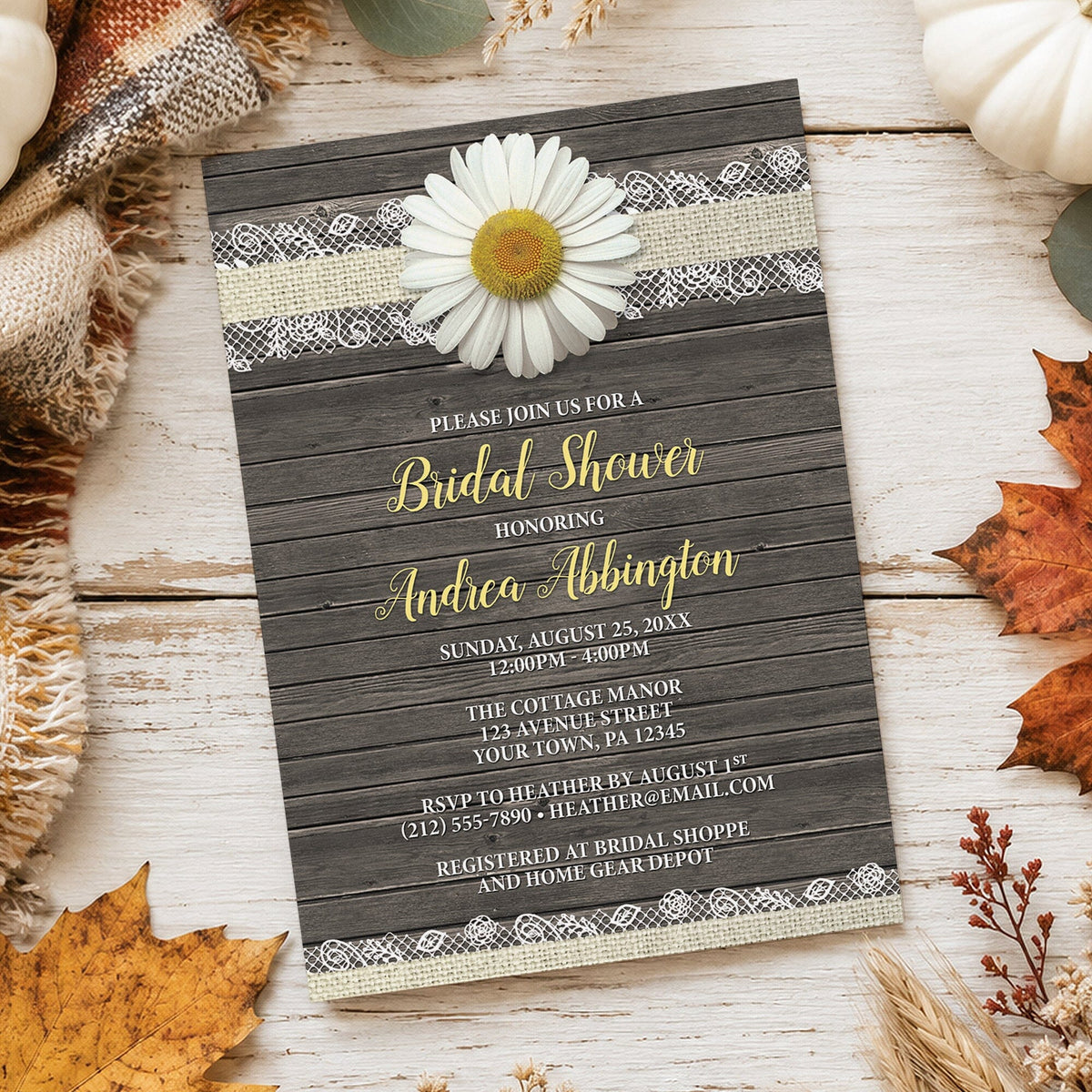 A rustic bridal shower invitation card styled on a whitewashed wood surface with autumn props. The card has a dark wood plank background, burlap and lace trim, a white daisy flower, and yellow and white text.