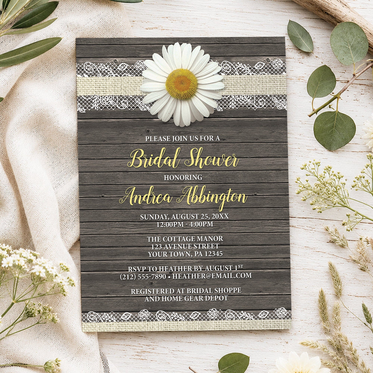 A rustic daisy bridal shower invitation styled in a flat-lay arrangement on a light wood surface with neutral fabric and greenery. The invitation has a dark wood plank background, a white daisy flower, and yellow and white text.