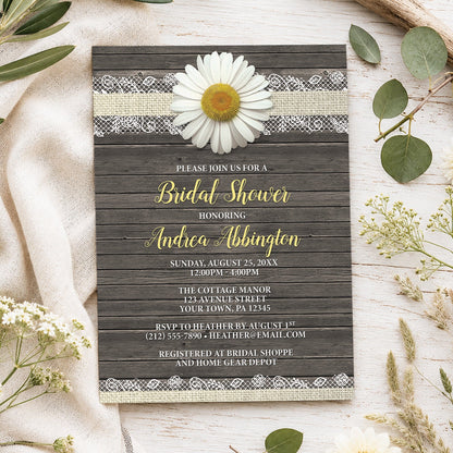 A rustic daisy bridal shower invitation styled in a flat-lay arrangement on a light wood surface with neutral fabric and greenery. The invitation has a dark wood plank background, a white daisy flower, and yellow and white text.