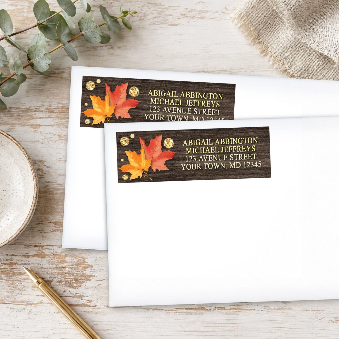 Falling Leaves with Gold Autumn Address Labels, shown on white envelopes, on a wooden surface with eucalyptus leaves and a pen.