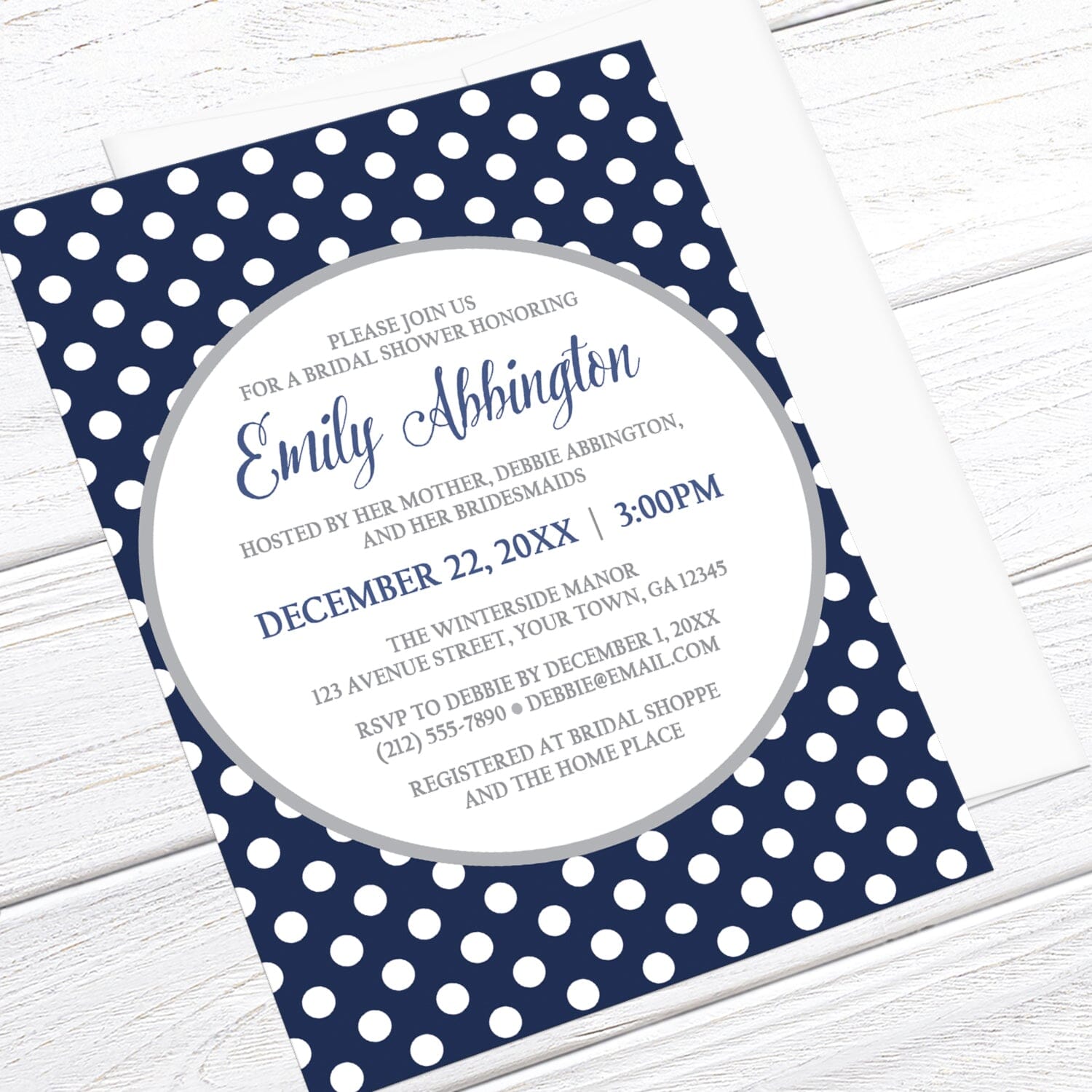 Navy blue and white polka dot bridal shower invitation shown at an angle on a light wooden surface with a white envelope.