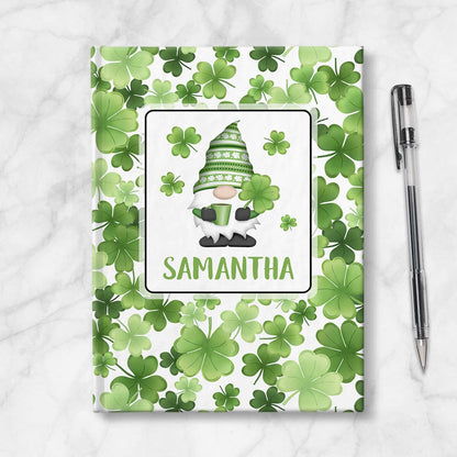 Personalized Green Shamrocks Gnome Journal at Artistically Invited. A custom book designed with an adorable gnome holding a hot beverage and a 4-leaf clover with 4 shamrocks floating around him in a white rectangular design on the front of the book, with your name personalized in green in a cute all capital letters font below the gnome. The background design that wraps around the cover of the book features a pattern of shamrocks and 4-leaf clovers in different shades of green. Book is next to a pen.
