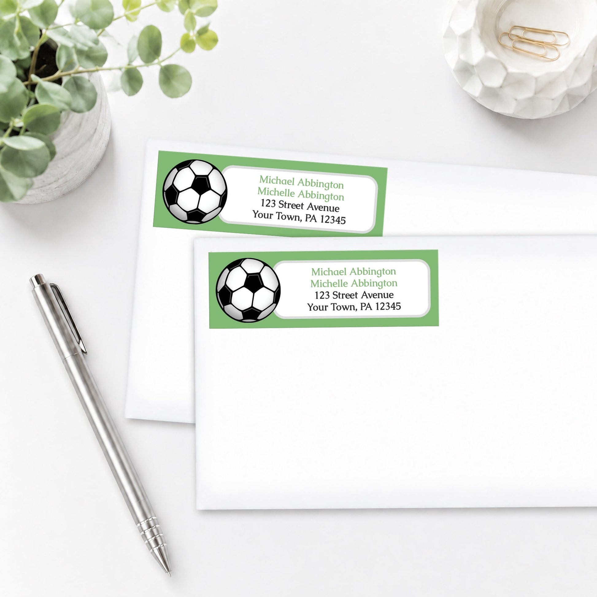 Green Soccer Ball Address Labels on white envelopes, shown on a white surface with a plant and a pen.