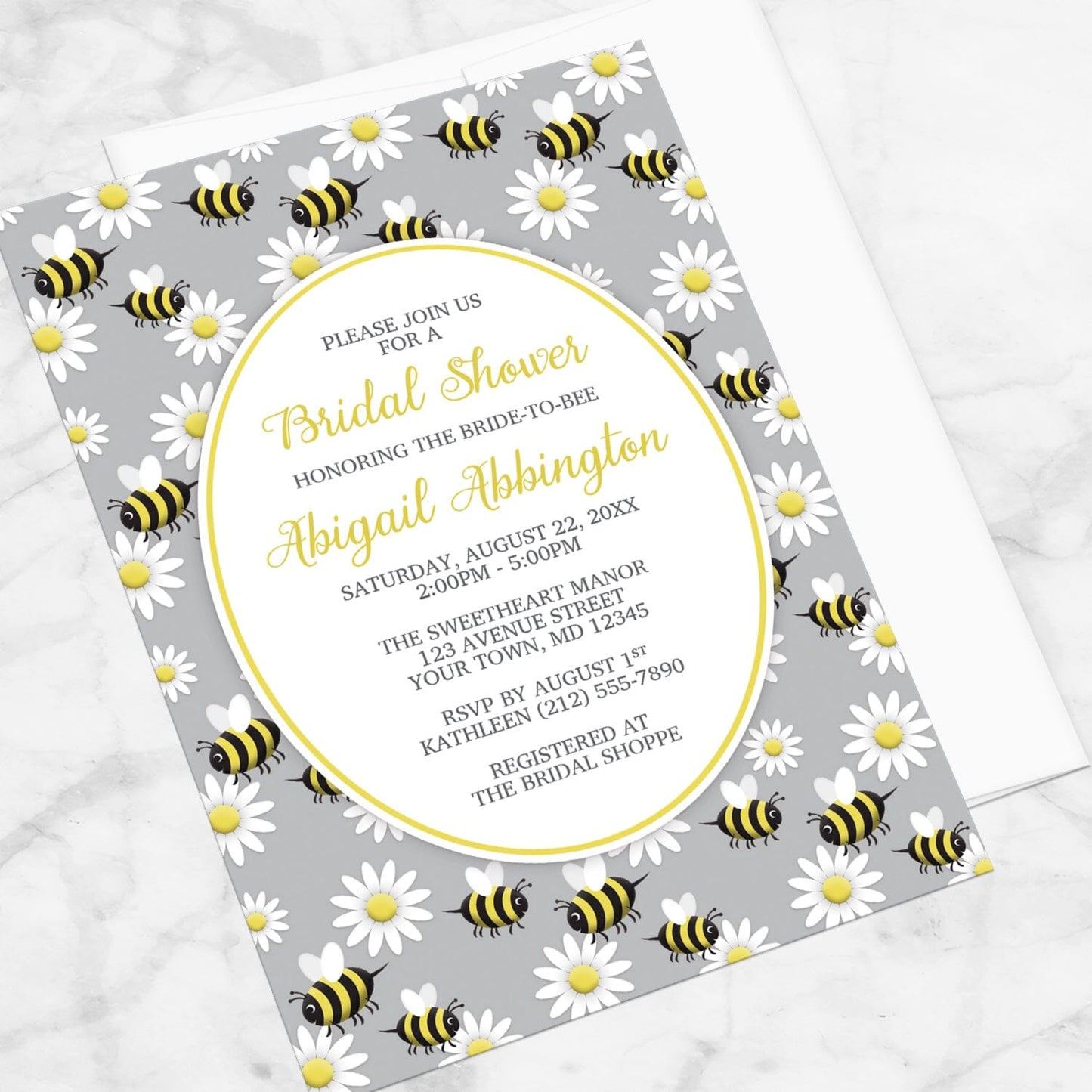 Bridal shower invitation with bee and daisy pattern on a gray background, displayed at an angle with a white envelope on a marble surface