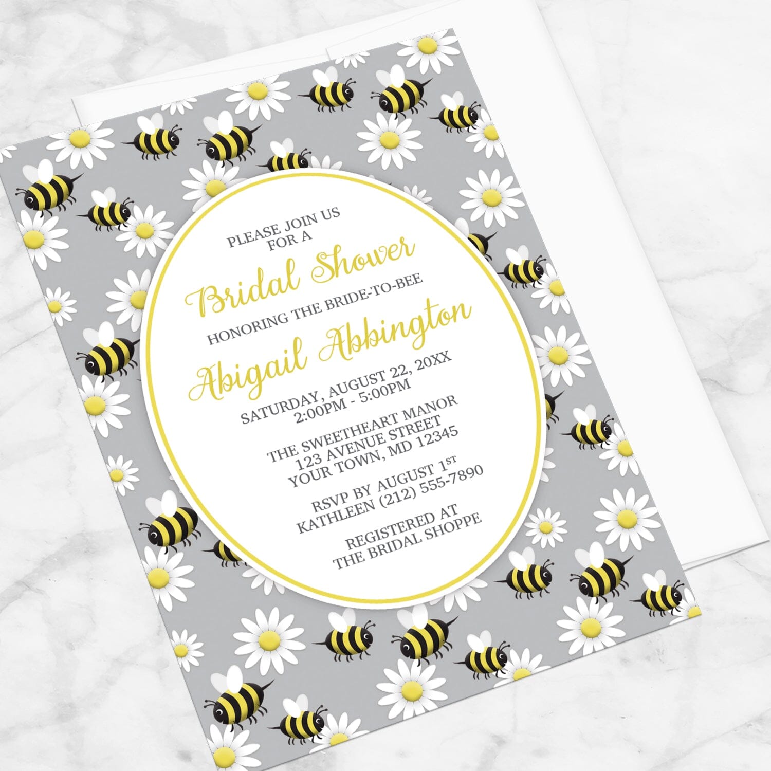 Bridal shower invitation with bee and daisy pattern on a gray background, displayed at an angle with a white envelope on a marble surface