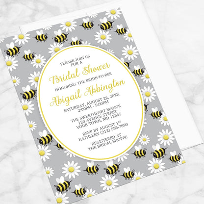 Bridal shower invitation with bee and daisy pattern on a gray background, displayed at an angle with a white envelope on a marble surface
