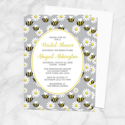 Bridal shower invitation with bee and daisy pattern on a gray background, displayed with a white envelope on a marble surface