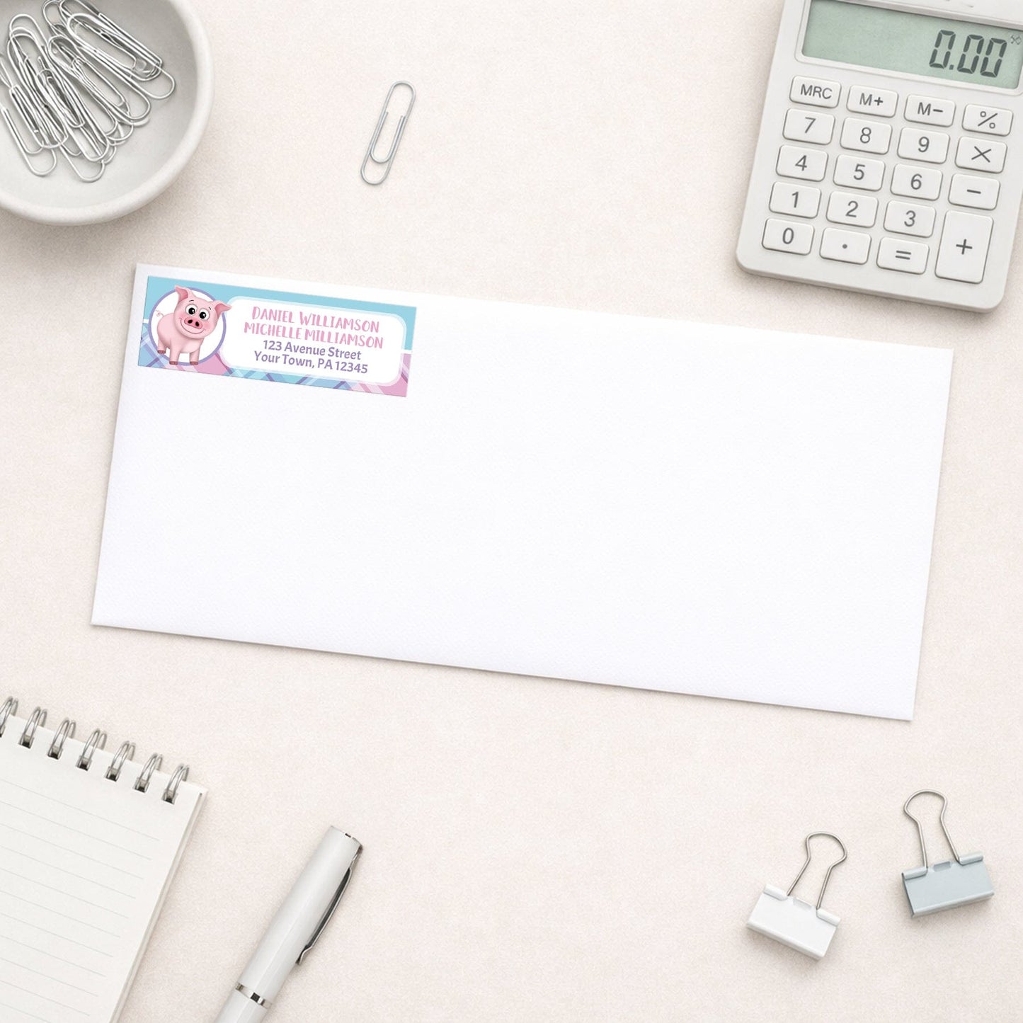 Happy Pig Pink Blue and Purple Plaid Address Label shown on a white envelope with everyday office supplies