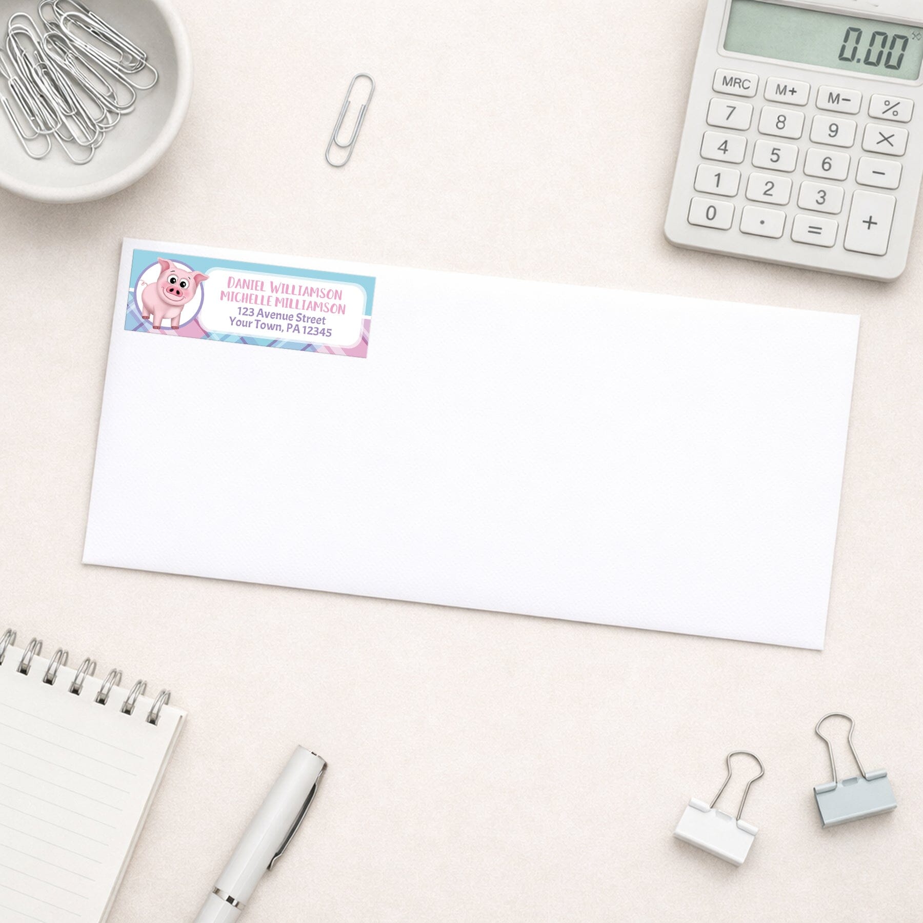 Happy Pig Pink Blue and Purple Plaid Address Label shown on a white envelope with everyday office supplies