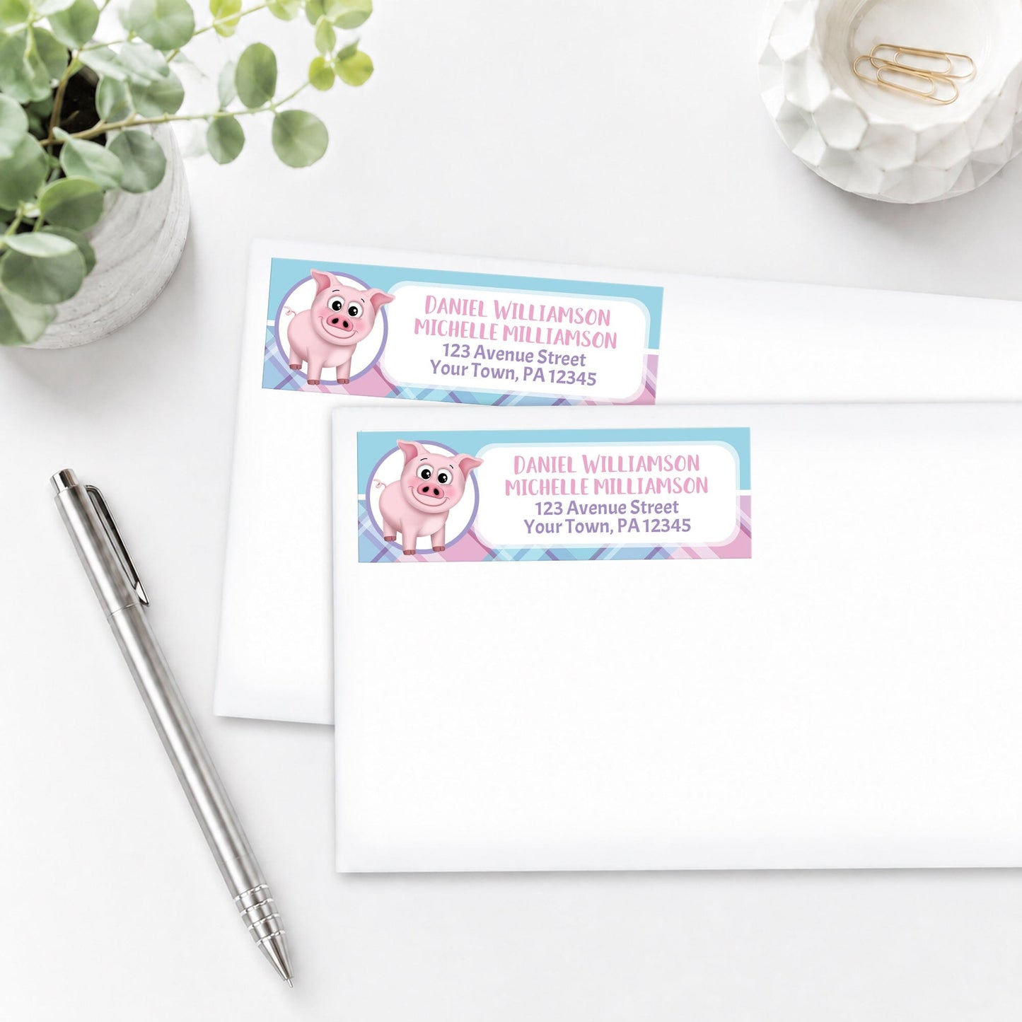 Happy Pig Pink Blue and Purple Plaid Address Labels, shown on white envelopes, on a white surface with a plant and a pen.