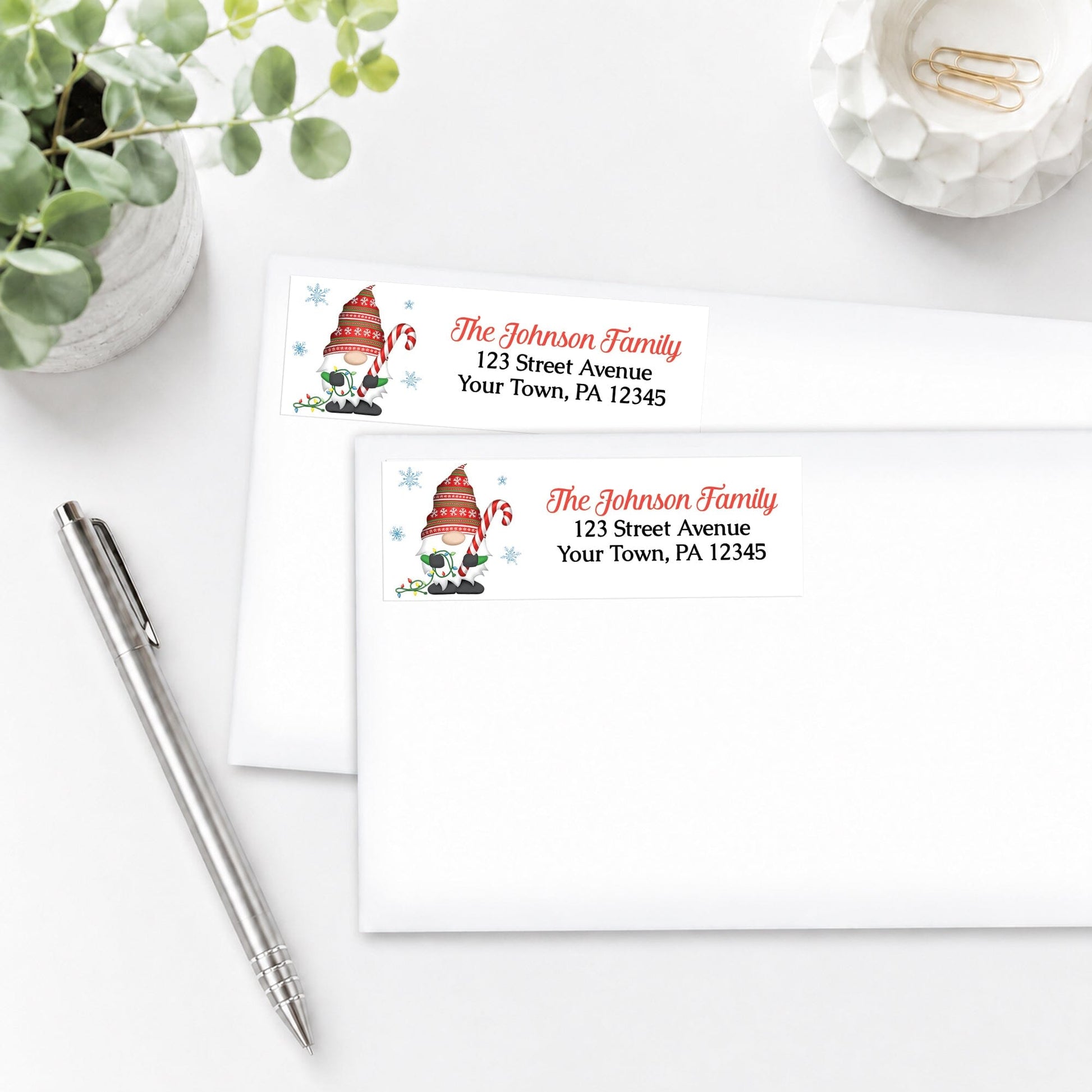 Two personalized Holiday Candy Cane Gnome Address Labels, shown on white envelopes, on a white surface with a pen and plant.