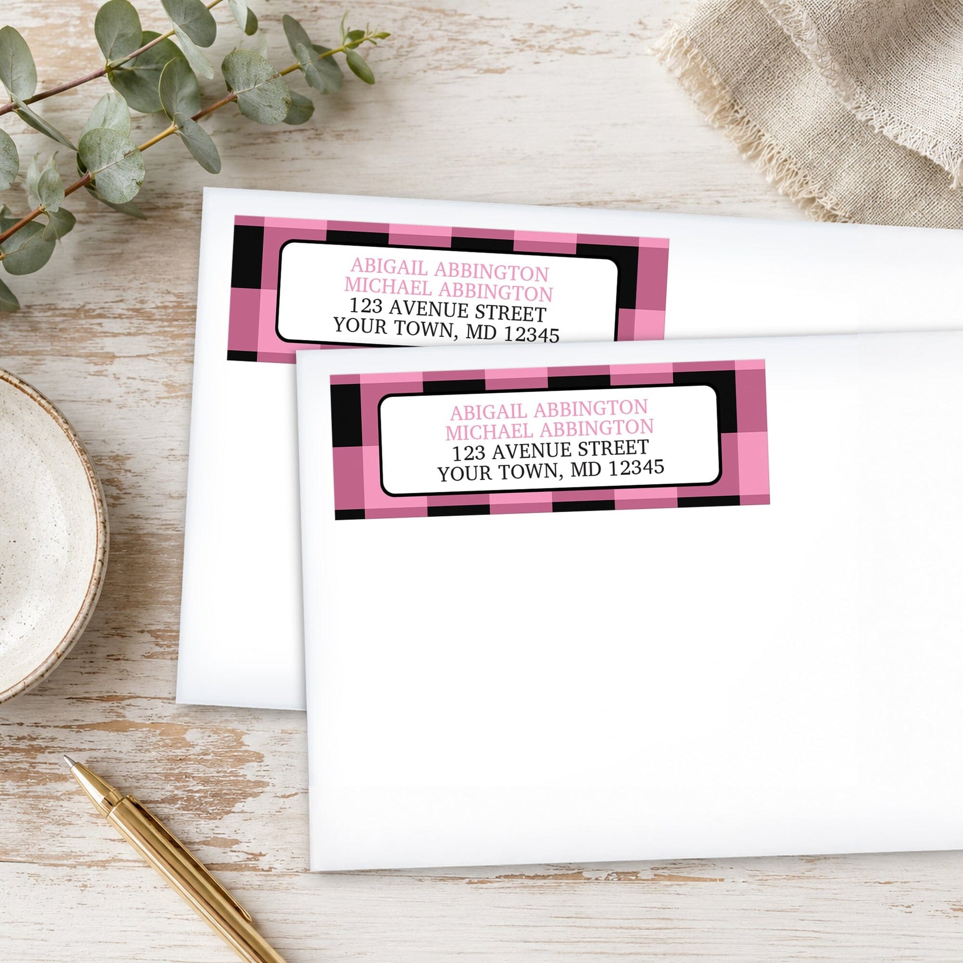 Light Pink and Black Buffalo Plaid Address Labels with pink and black checkered design, shown on white envelopes, on a wooden surface with eucalyptus leaves and a pen.