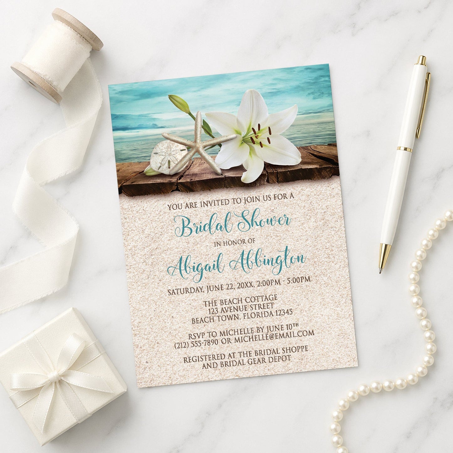 Lily Seashells and Sand Beach Bridal Shower Invitation on a marble surface with white ribbon, gift box, pen, and string of pearls beside it.