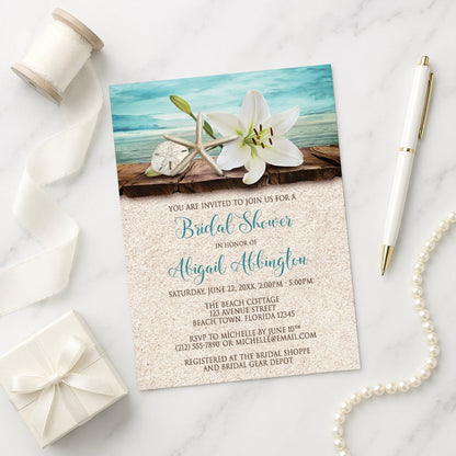 Lily Seashells and Sand Beach Bridal Shower Invitation on a marble surface with white ribbon, gift box, pen, and string of pearls beside it.