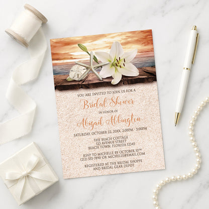 Lily Seashells and Sand Autumn Beach Bridal Shower Invitation on a marble surface with ribbon and pen.