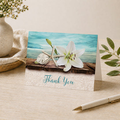 Lily Seashells and Sand Beach Thank You Card shown on a tabletop with greenery, a vase, and a pen.
