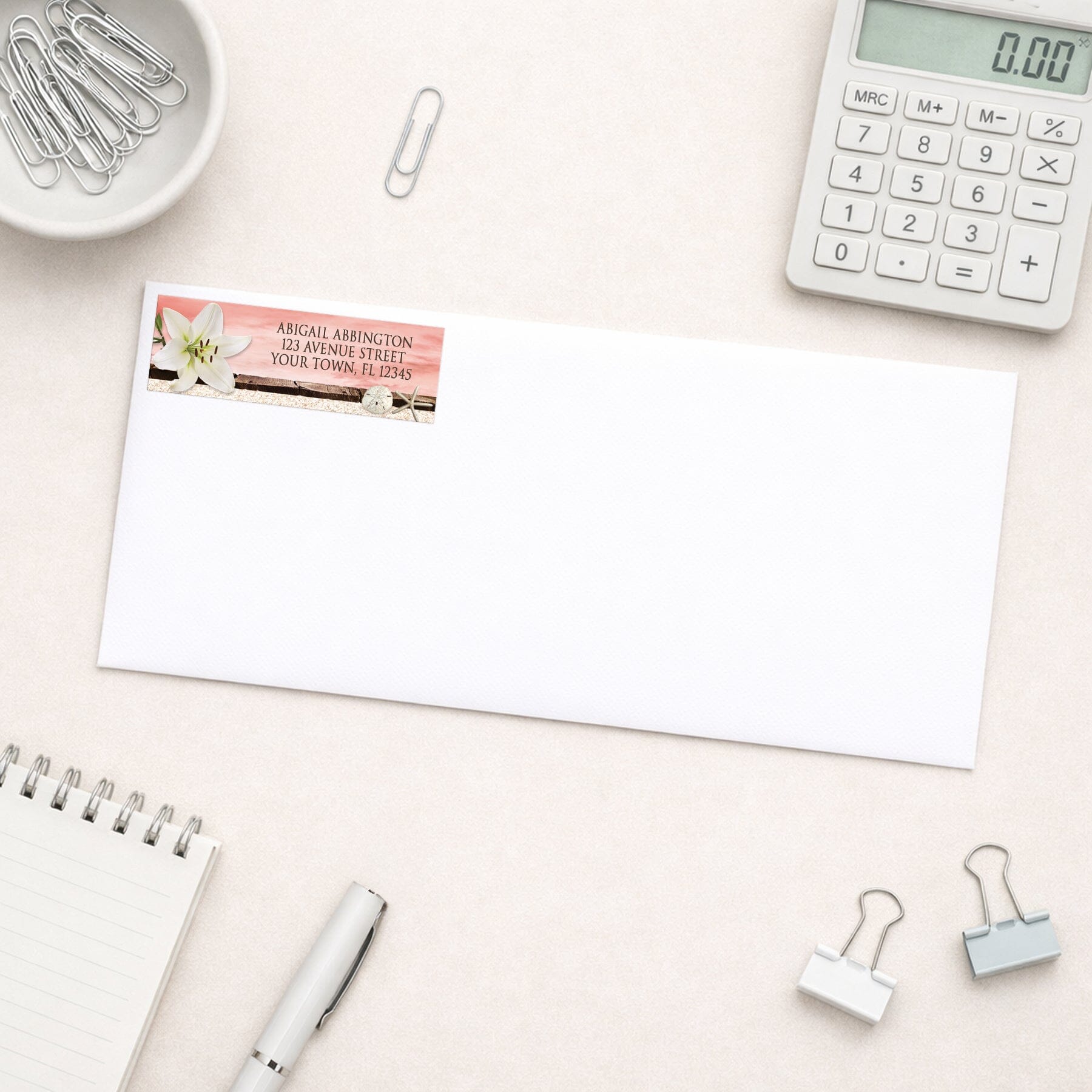 Lily Seashells and Sand Coral Beach Address Label shown on a white envelope with everyday office supplies