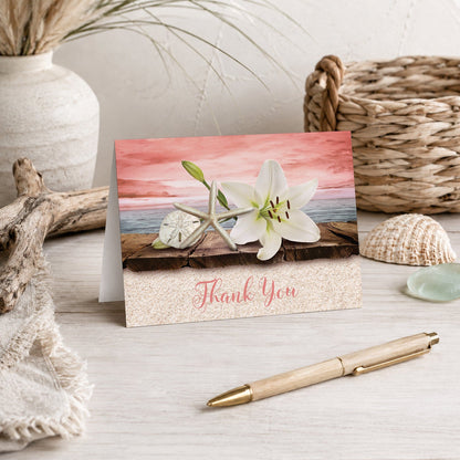 Lily Seashells and Sand Coral Beach Thank You Card shown on a beach house tabletop with coastal decor and a pen.
