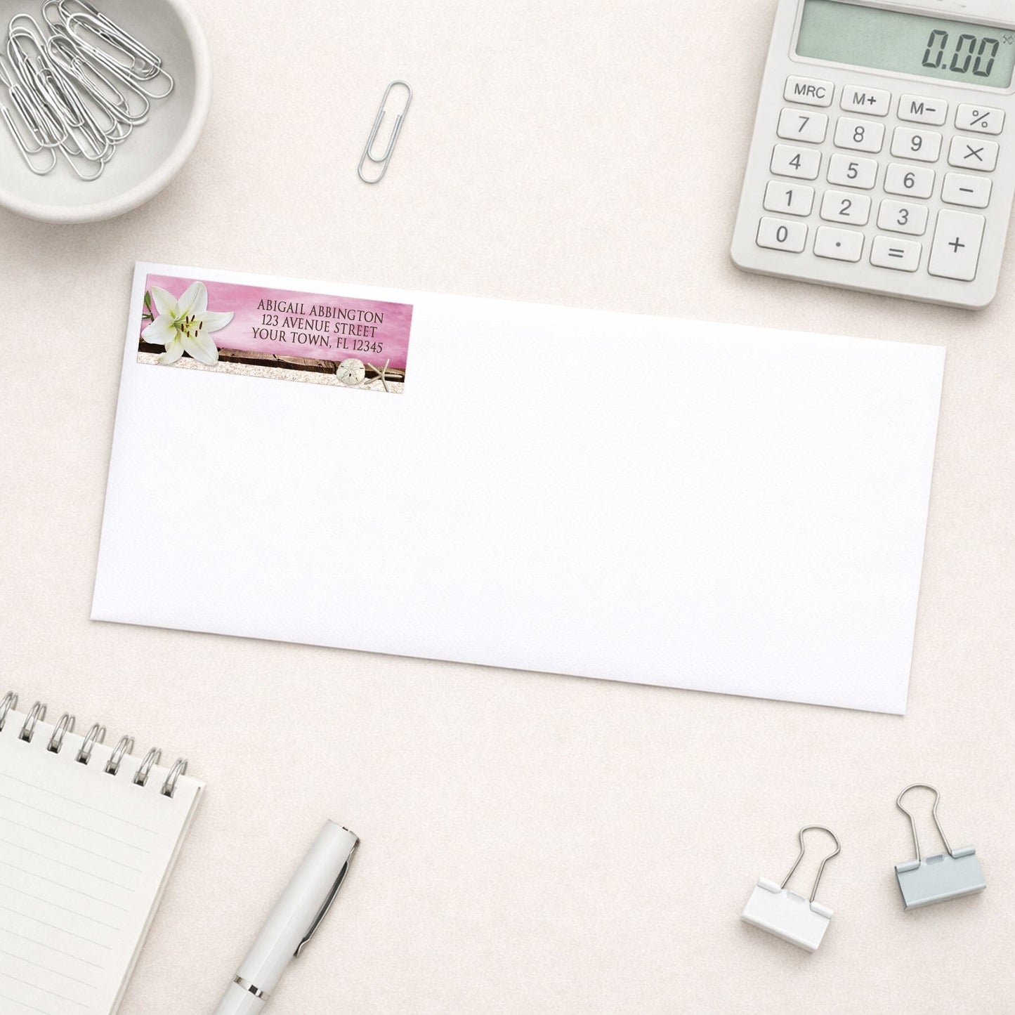 Lily Seashells and Sand Magenta Beach Address Label shown on a white envelope with everyday office supplies
