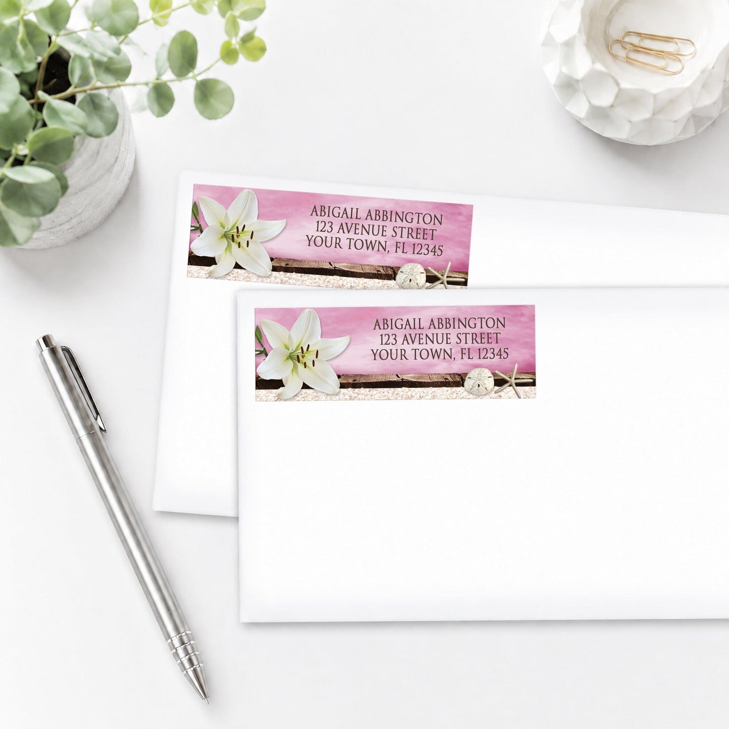 Lily Seashells and Sand Magenta Beach Address Labels shown on white envelopes, on a white surface with a plant and pen.
