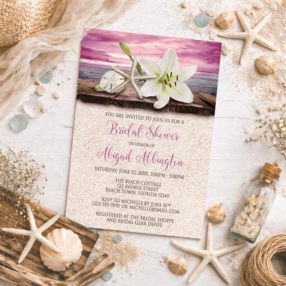 Lily Seashells and Sand Magenta Beach Bridal Shower Invitation on a beachy surface with sand, seashells, drift wood, and other beach-themed decor.