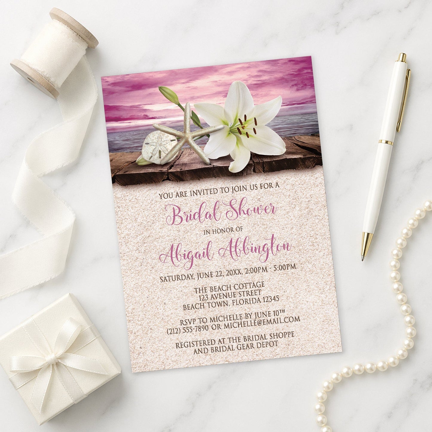 Lily Seashells and Sand Magenta Beach Bridal Shower Invitation on a marble surface with a white gift box, pen, ribbon, and pearls.