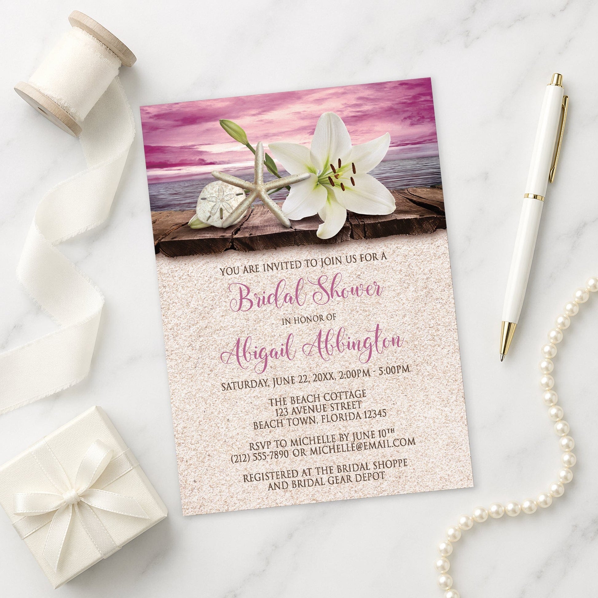 Lily Seashells and Sand Magenta Beach Bridal Shower Invitation on a marble surface with a white gift box, pen, ribbon, and pearls.