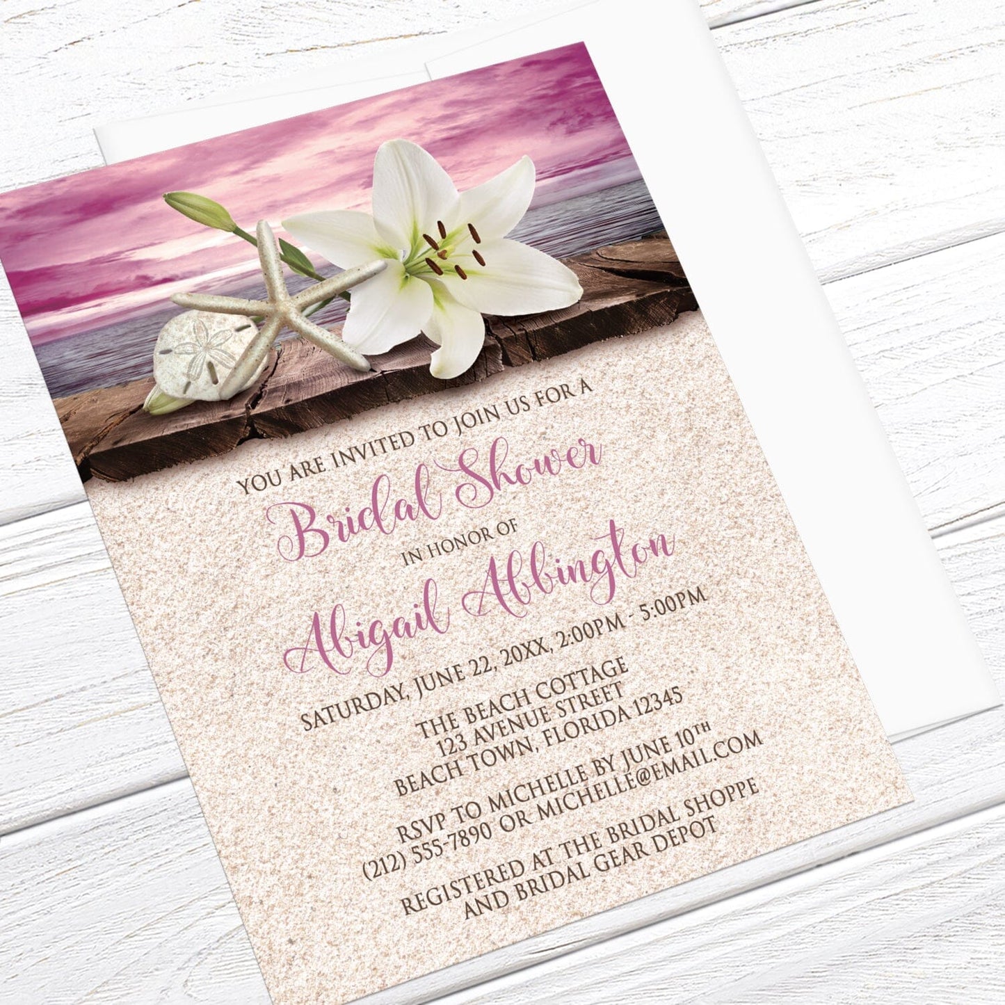Lily Seashells and Sand Magenta Beach Bridal Shower Invitation shown at an angle on a light wood surface with a white envelope.