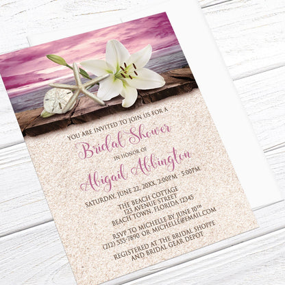 Lily Seashells and Sand Magenta Beach Bridal Shower Invitation shown at an angle on a light wood surface with a white envelope.