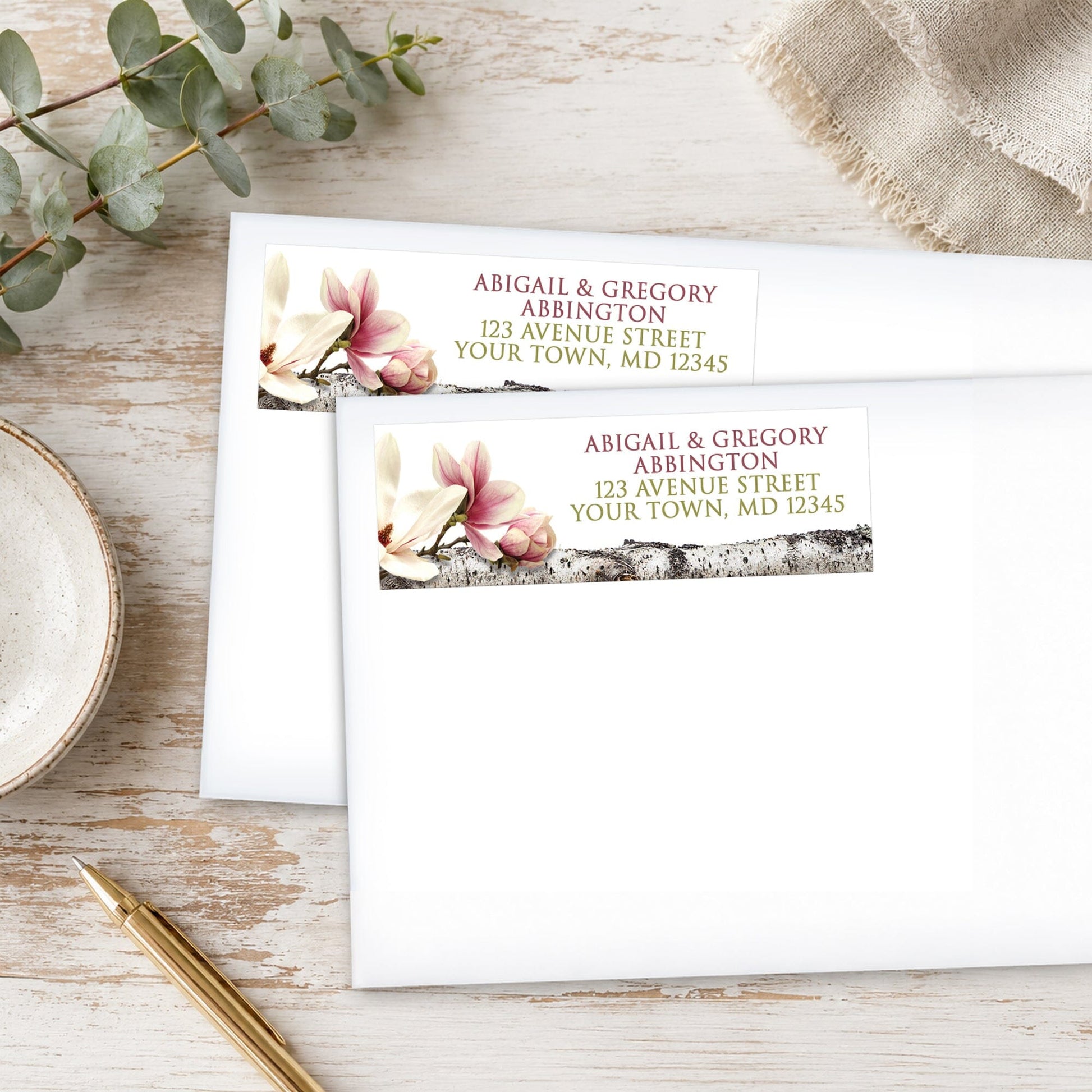 Address labels with magnolia and birch design, shown on white envelopes, on a wooden surface with eucalyptus leaves and a pen.