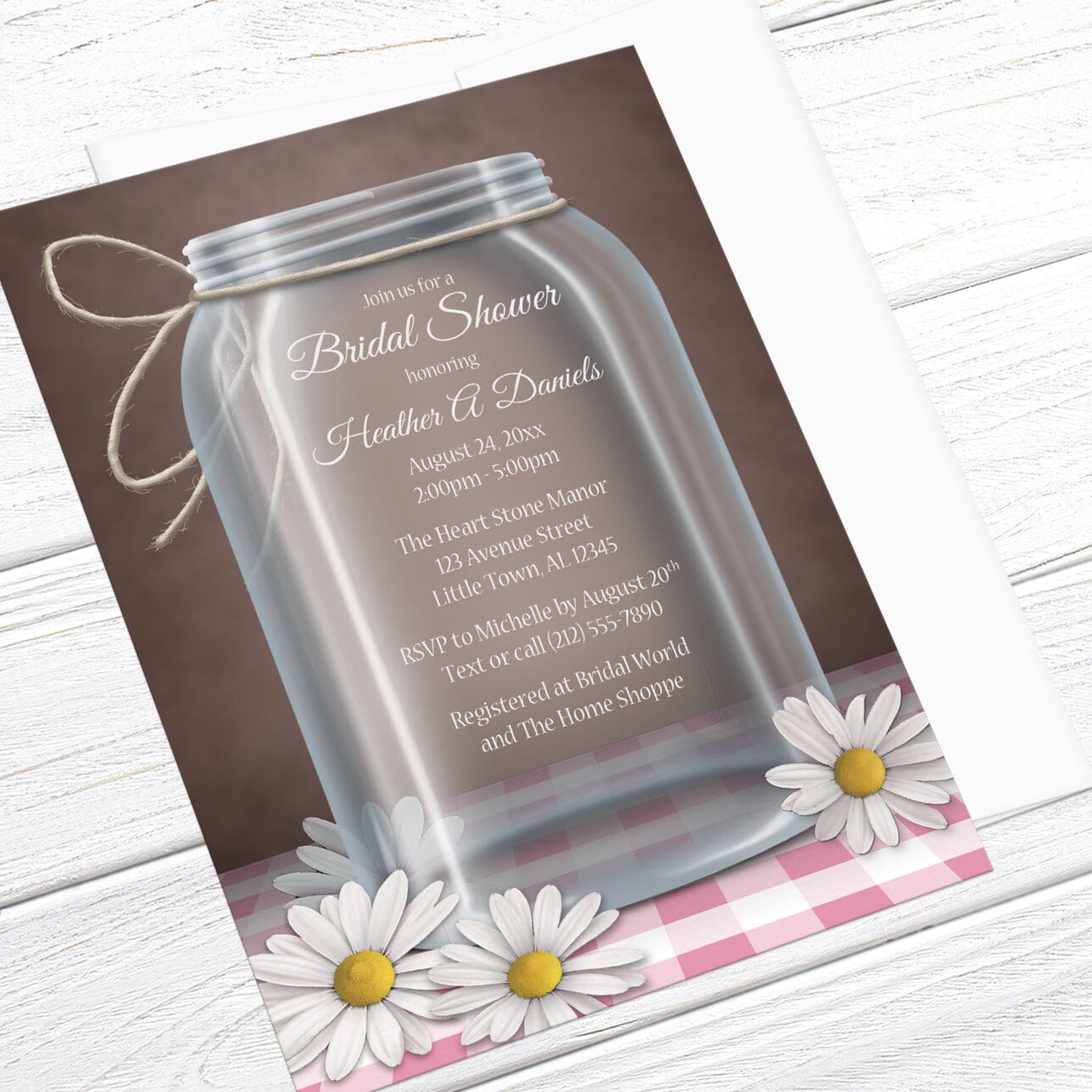 Bridal Shower invitation with a mason jar design with white daisies and pink gingham over a rustic brown background, displayed at an angle with a white envelope over a white wood surface
