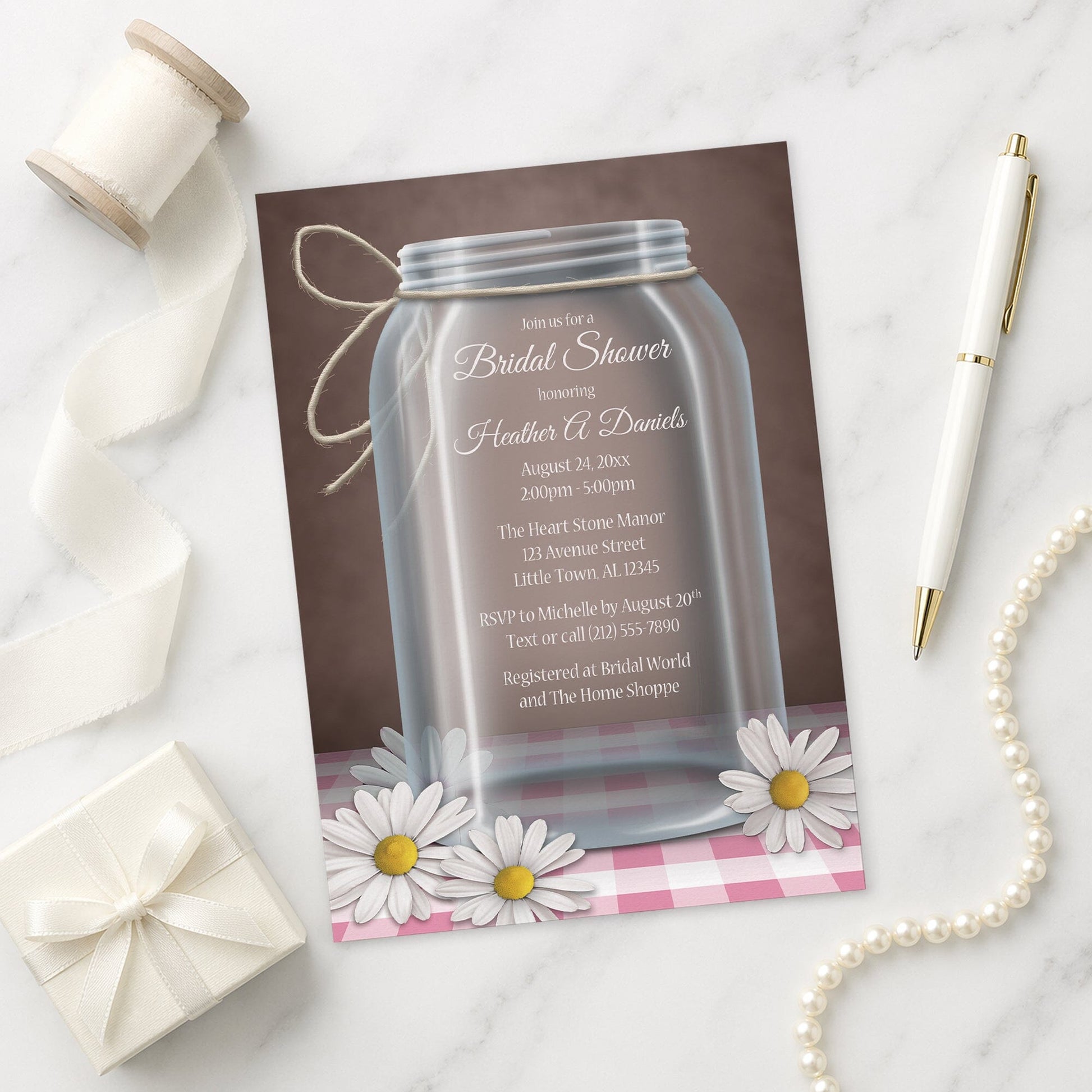 Mason Jar Daisy Pink Gingham Bridal Shower Invitation on a marble surface with decorative items.