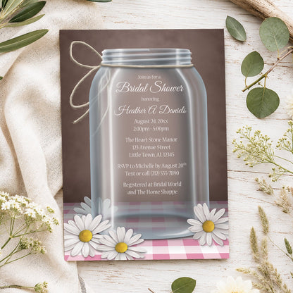 Country mason jar daisy gingham bridal shower invitations with an illustration of a glass mason jar with twine tied around the neck of it, and white and yellow daisies laying at the base of the jar on a pink gingham tabletop and over a vintage brown background, styled on a light rustic surface with spring and summer-inspired plants and beige fabric.