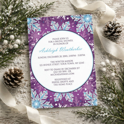 Modern Purple Blue Snowflake Bridal Shower Invitation shown on a rustic wooden surface with winter greenery, pine cones, and ribbon.