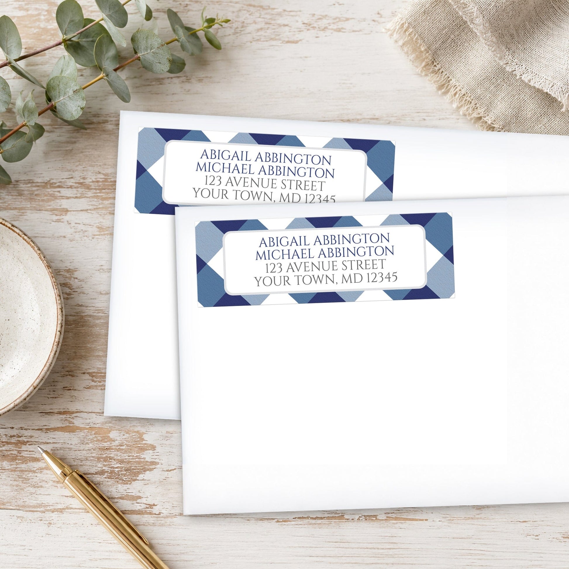 Two Navy Blue Gingham Address Labels on white envelopes shown with a pen and eucalyptus branch on a wooden surface.