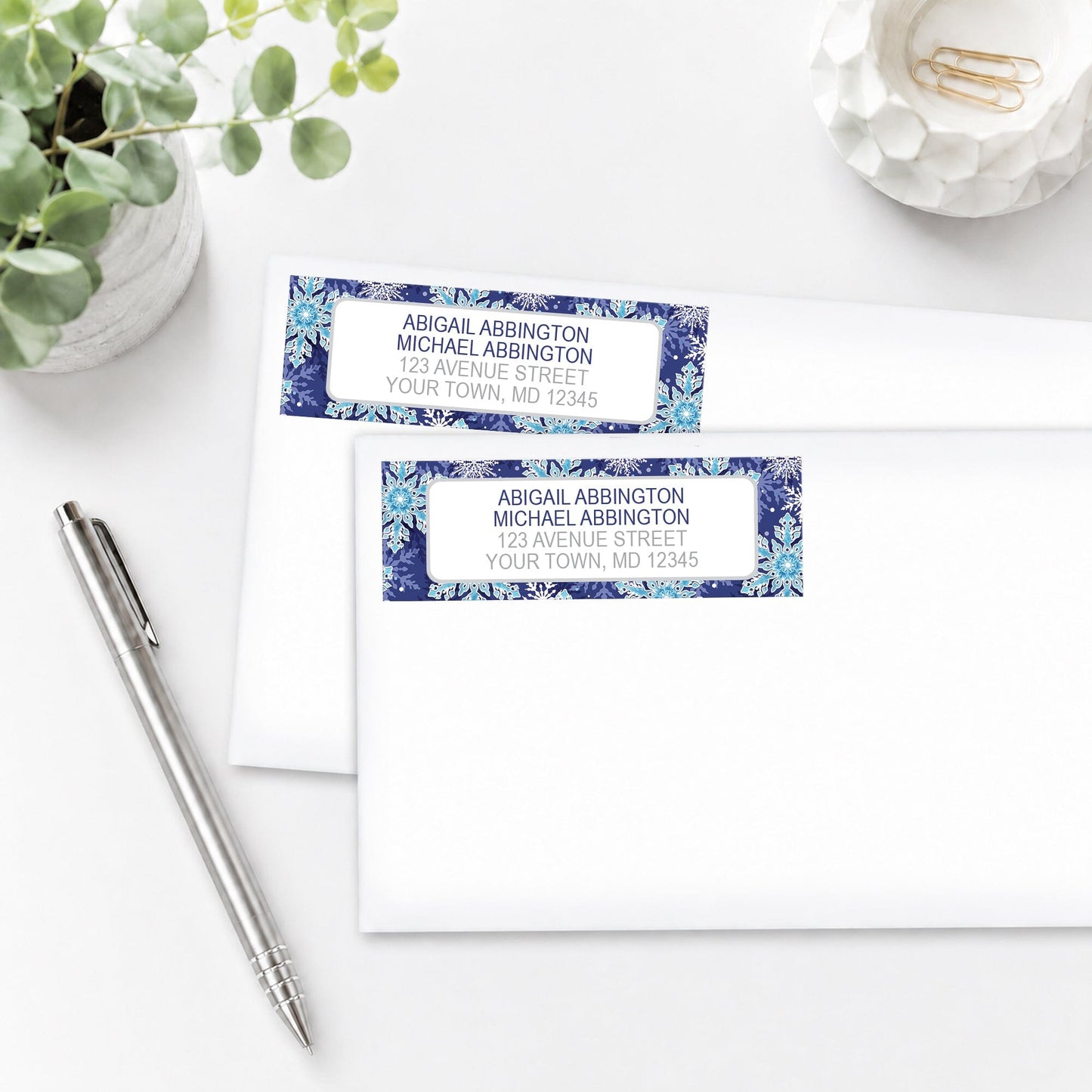 Navy Blue Snowflake Winter Address Labels on white envelopes with a pen and plant in the background.