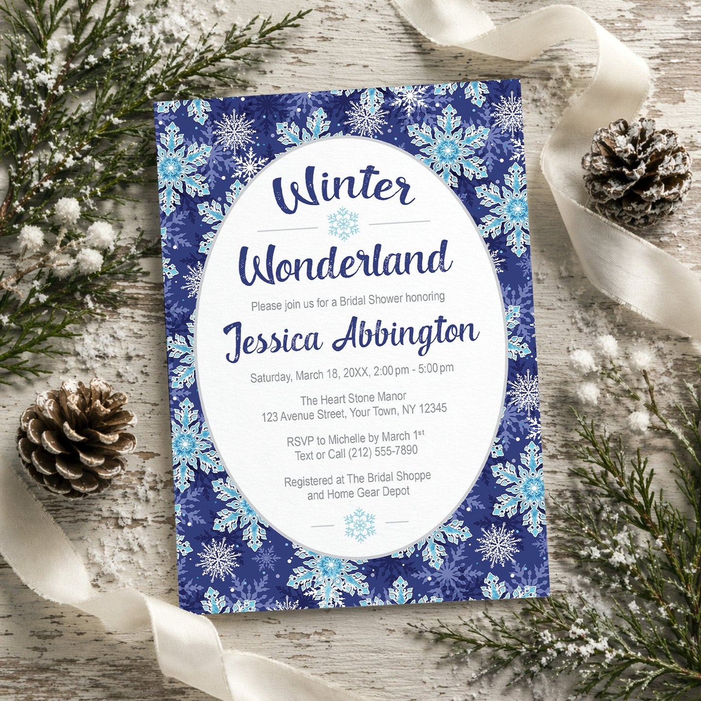 Winter Wonderland bridal shower invitation with a navy blue snowflake design shown on a wooden surface with pine cones and ribbons.