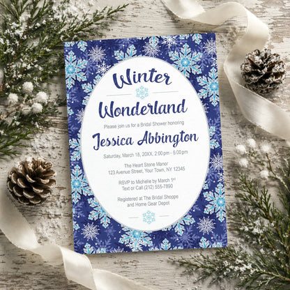 Winter Wonderland bridal shower invitation with a navy blue snowflake design shown on a wooden surface with pine cones and ribbons.