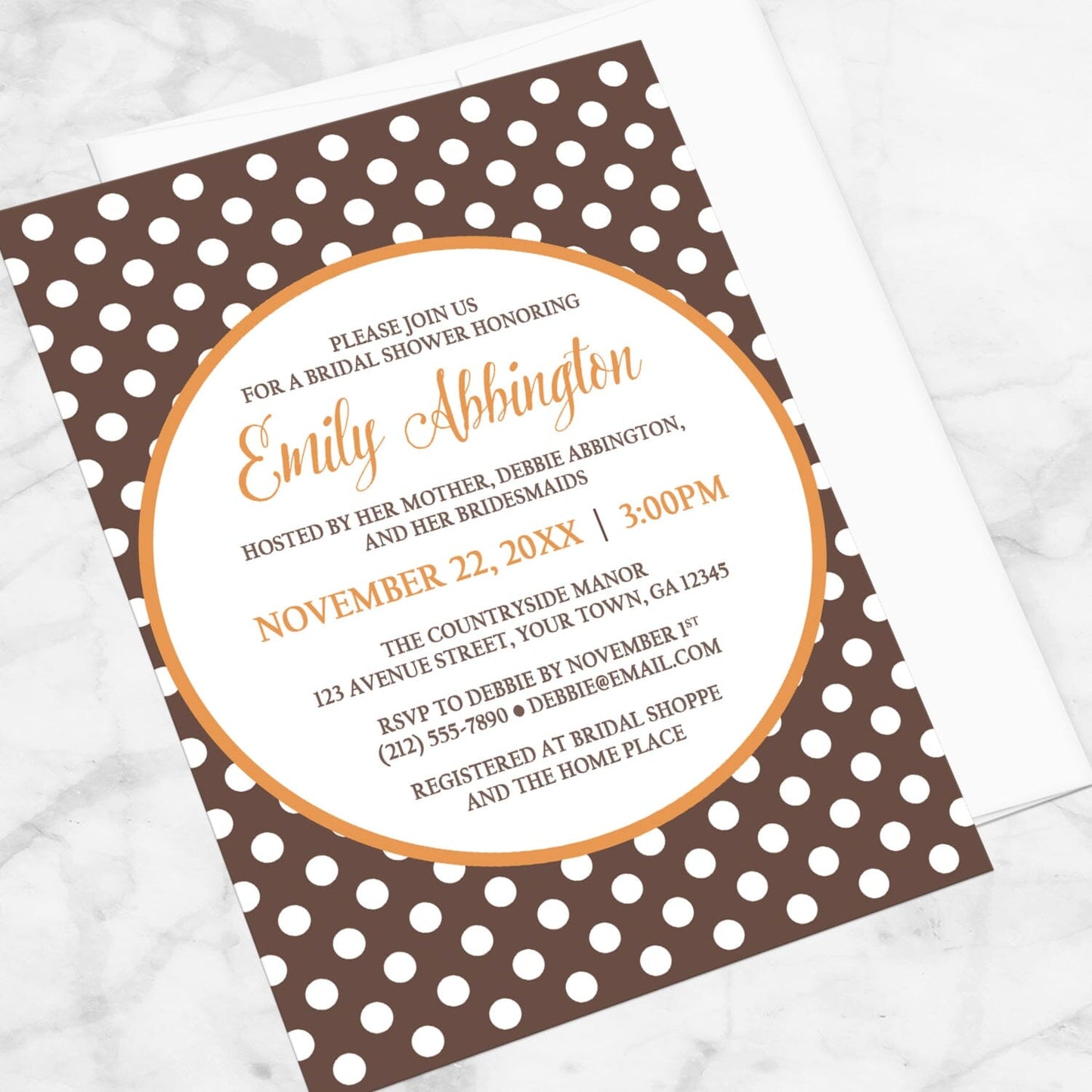 Bridal Shower invitation with a brown polka dot pattern and orange accents, displayed at an angle with a white envelope on a marble surface