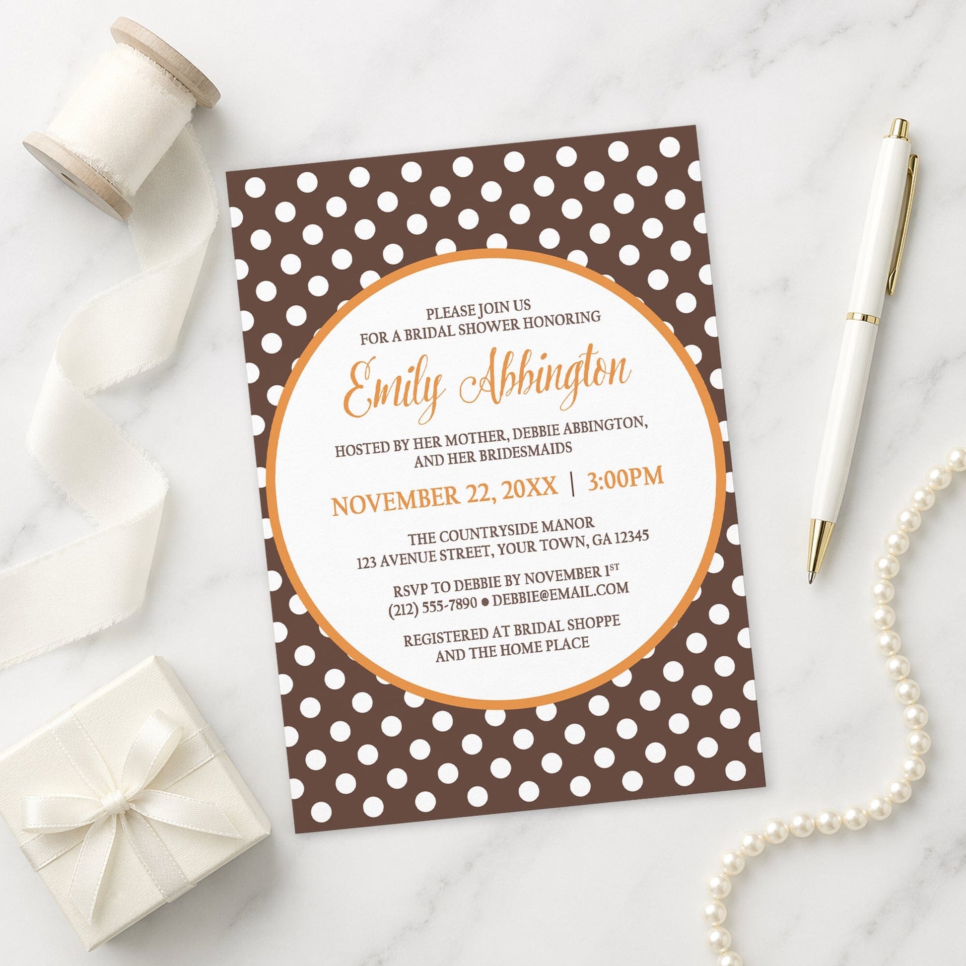Orange Brown Polka Dot Bridal Shower Invitation on a marble surface with a white gift box, ribbon, pen, and pearls.