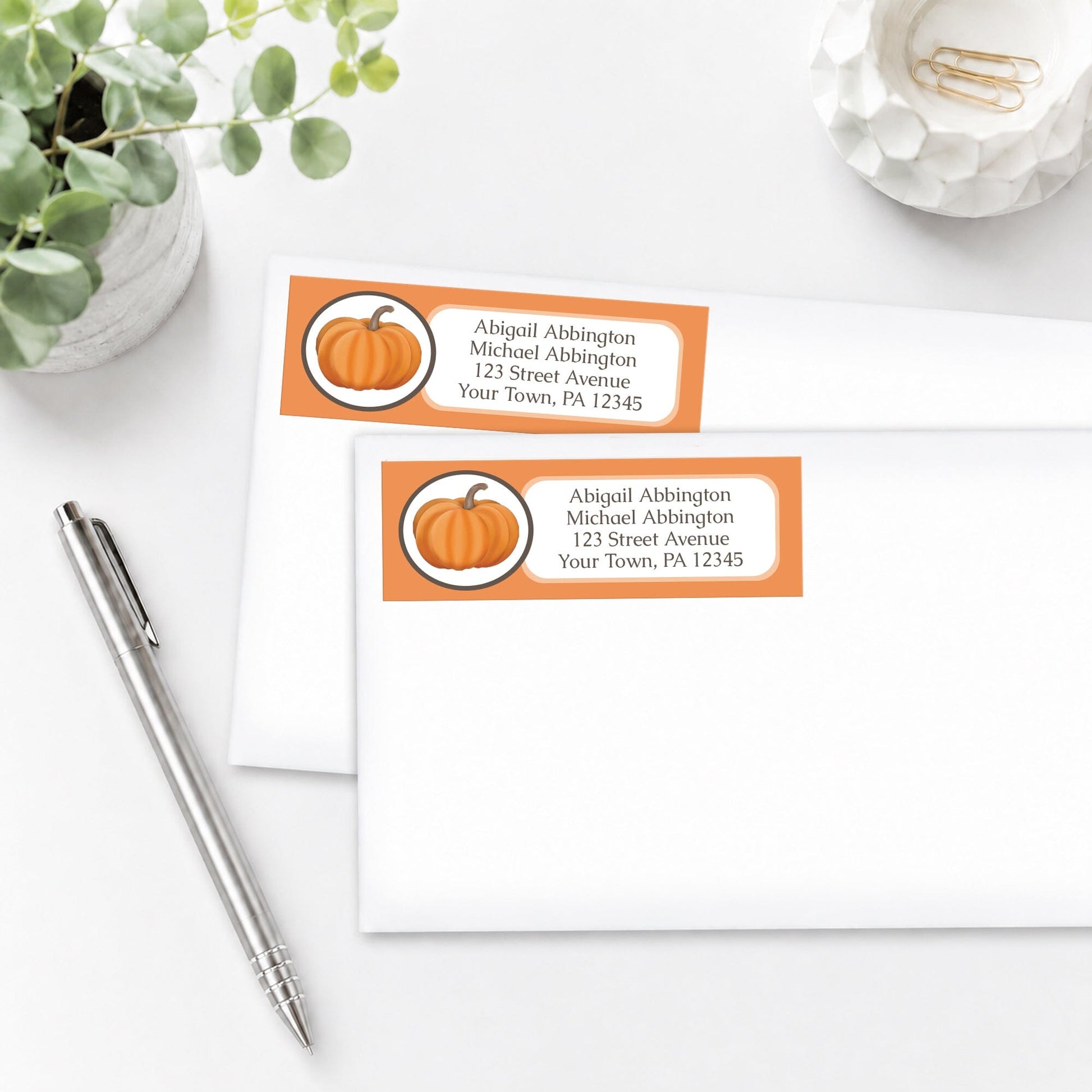 Orange Pumpkin Address Labels shown on white envelopes, on a white surface with a plant and a pen.