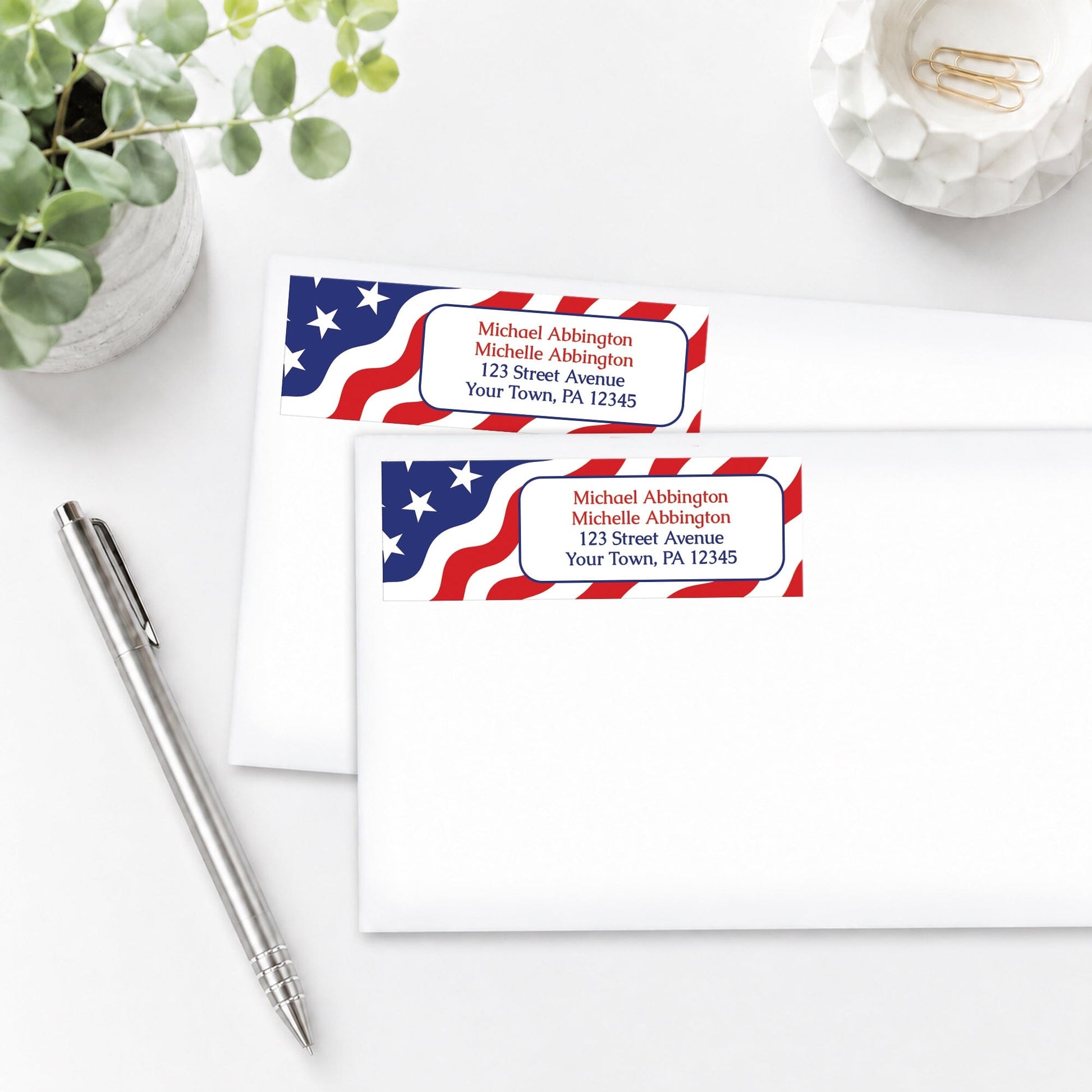 Two address labels with American flag design shown on envelopes on a white surface with a pen and plant.