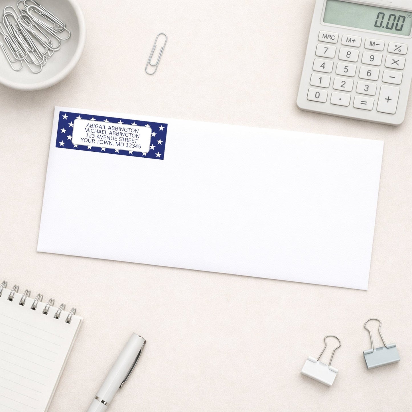 White envelope with Patriotic White Stars on Blue Address Label on a light surface with office supplies.