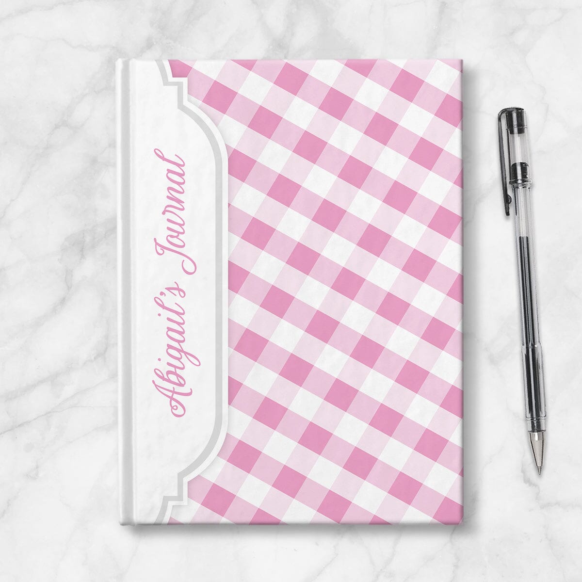 Personalized Pink Gingham Journal at Artistically Invited. Image shows the book on a countertop next to a pen.