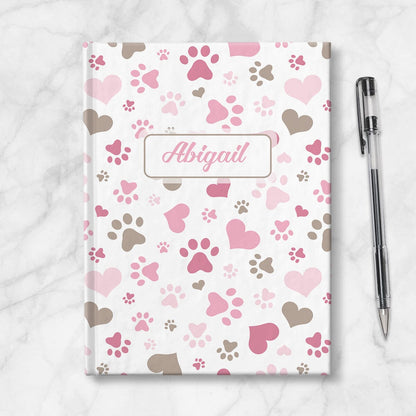 Personalized Pink Hearts and Paw Prints Journal at Artistically Invited. Image shows the book on a countertop next to a pen.